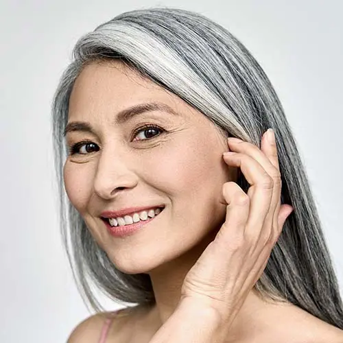 A woman with gray hair is smiling and holding her hand to the ear.